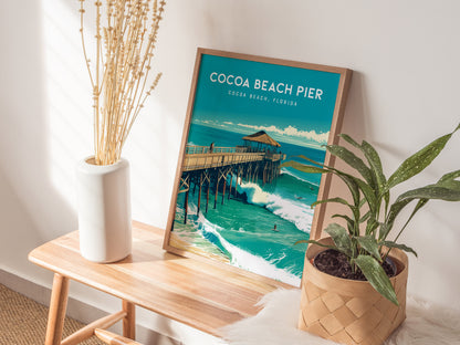 A framed Cocoa Beach Pier poster rests on a wooden table beside a white vase with dried flowers and a potted plant, illuminated by sunlight.