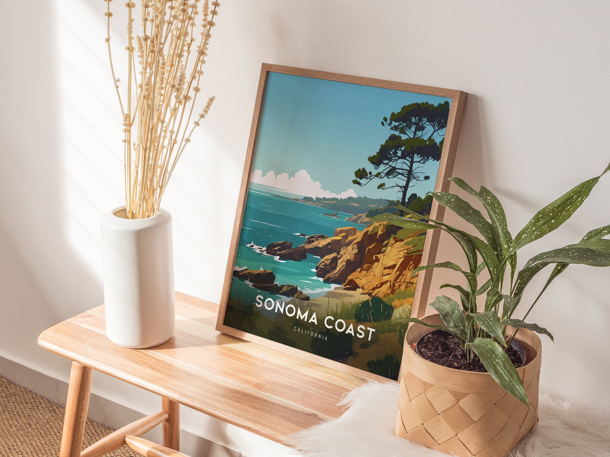 A framed Sonoma Coast poster rests on a wooden bench beside a white vase with dried flowers and a green plant in a woven basket.