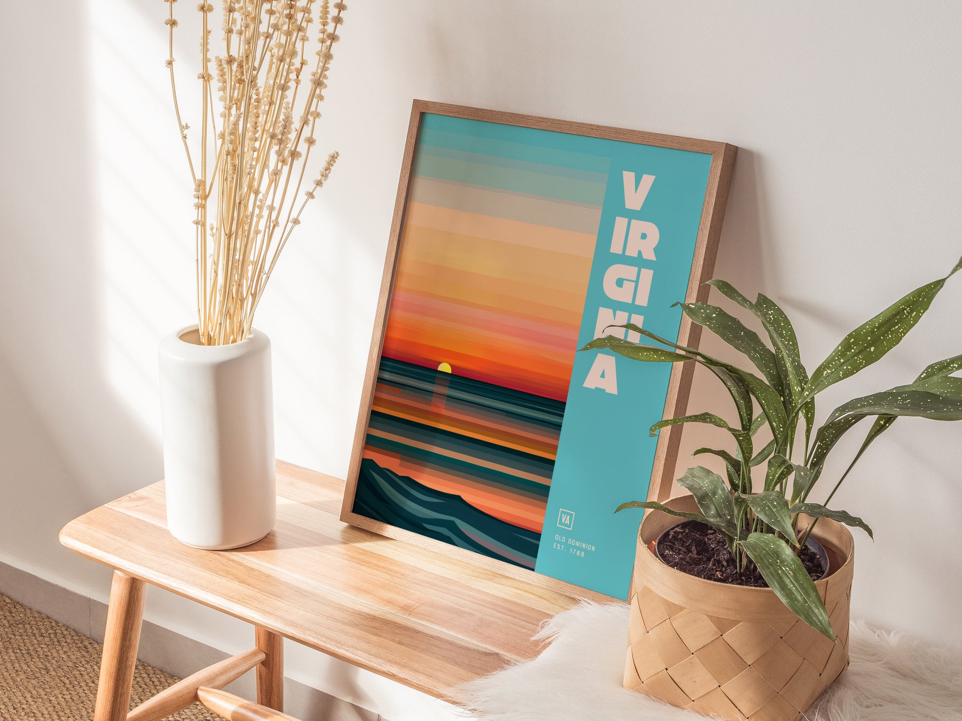 A framed Virginia poster with vibrant sunset colors and bold text rests on a wooden bench, accompanied by a white vase with dried flowers and a potted plant.