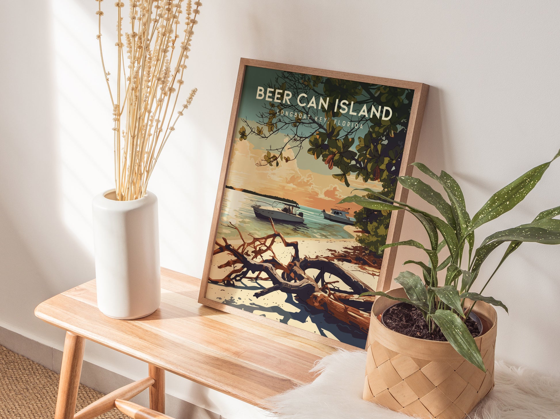 A framed poster of Beer Can Island, Longboat Key, depicting boats and driftwood on a beach, rests on a wooden bench. Nearby, a white vase with dried flowers.
