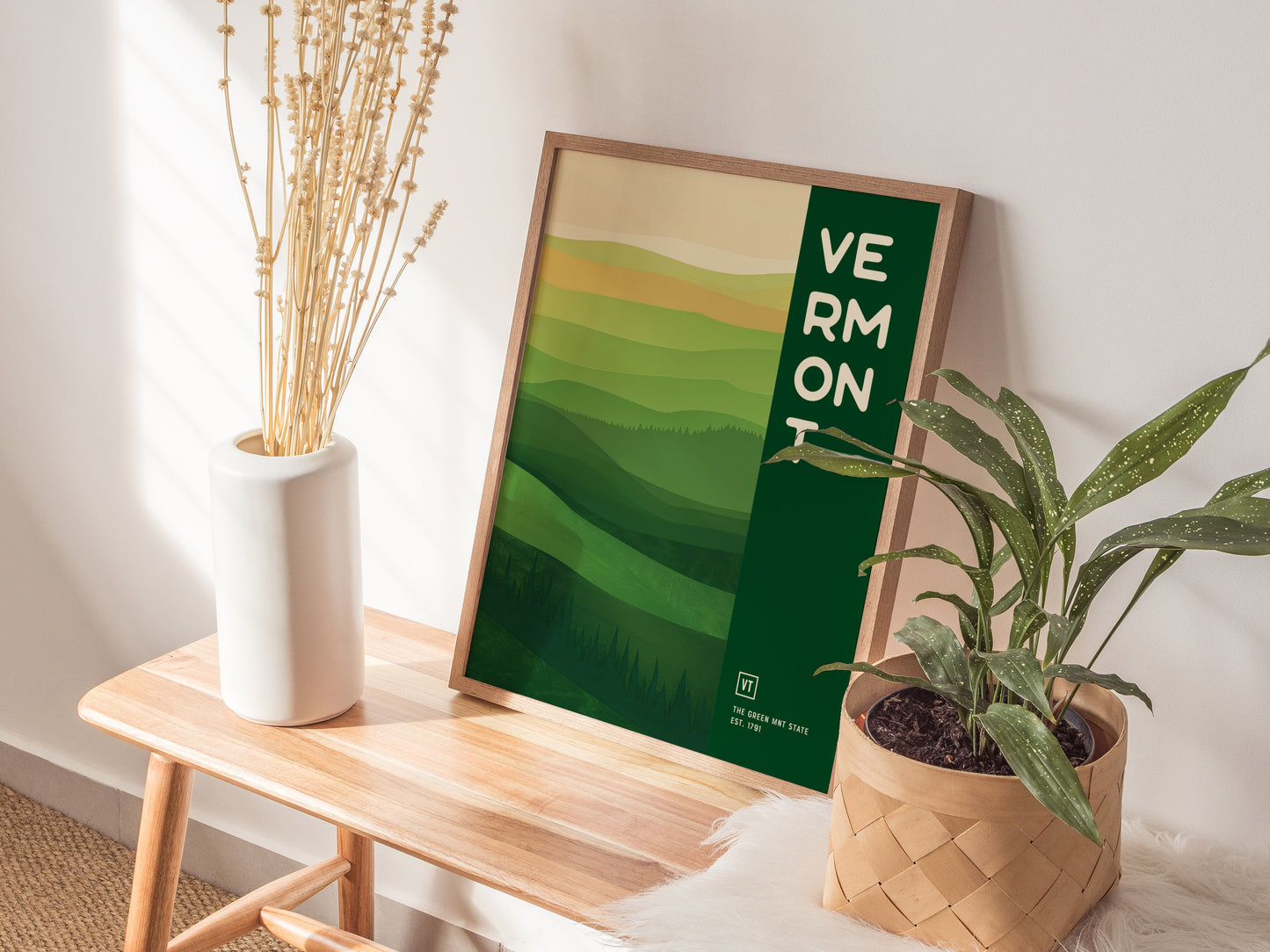 A framed Vermont poster with abstract green hills is propped against a wall on a wooden bench. Nearby, a white vase with dried flowers.