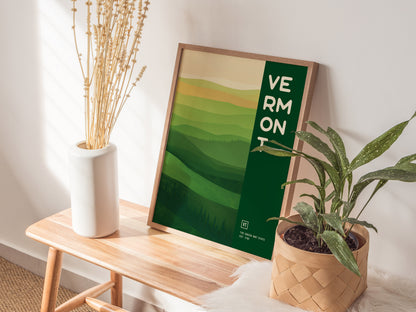 A framed Vermont poster with abstract green hills is propped against a wall on a wooden bench. Nearby, a white vase with dried flowers.