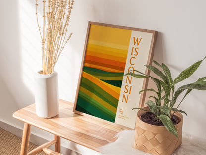 A minimalist Wisconsin poster with green and gold abstract layers sits on a wooden bench, alongside a white vase with dried flowers and a potted plant.