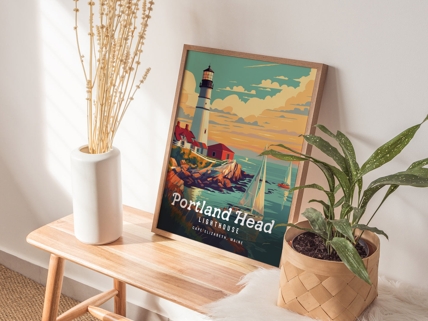 A framed Portland Head Lighthouse poster rests on a wooden bench beside a white vase with dried flowers and a potted plant. Sunlight casts shadows on the wall.