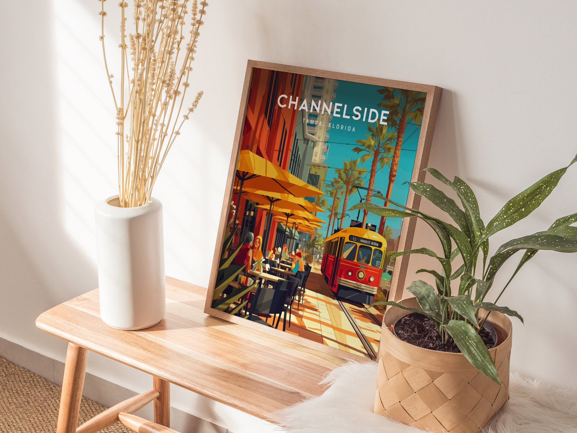 A framed Channelside Tampa poster sits on a wooden bench beside a vase of dried flowers and a potted plant, depicting a vibrant street scene with a yellow and red.