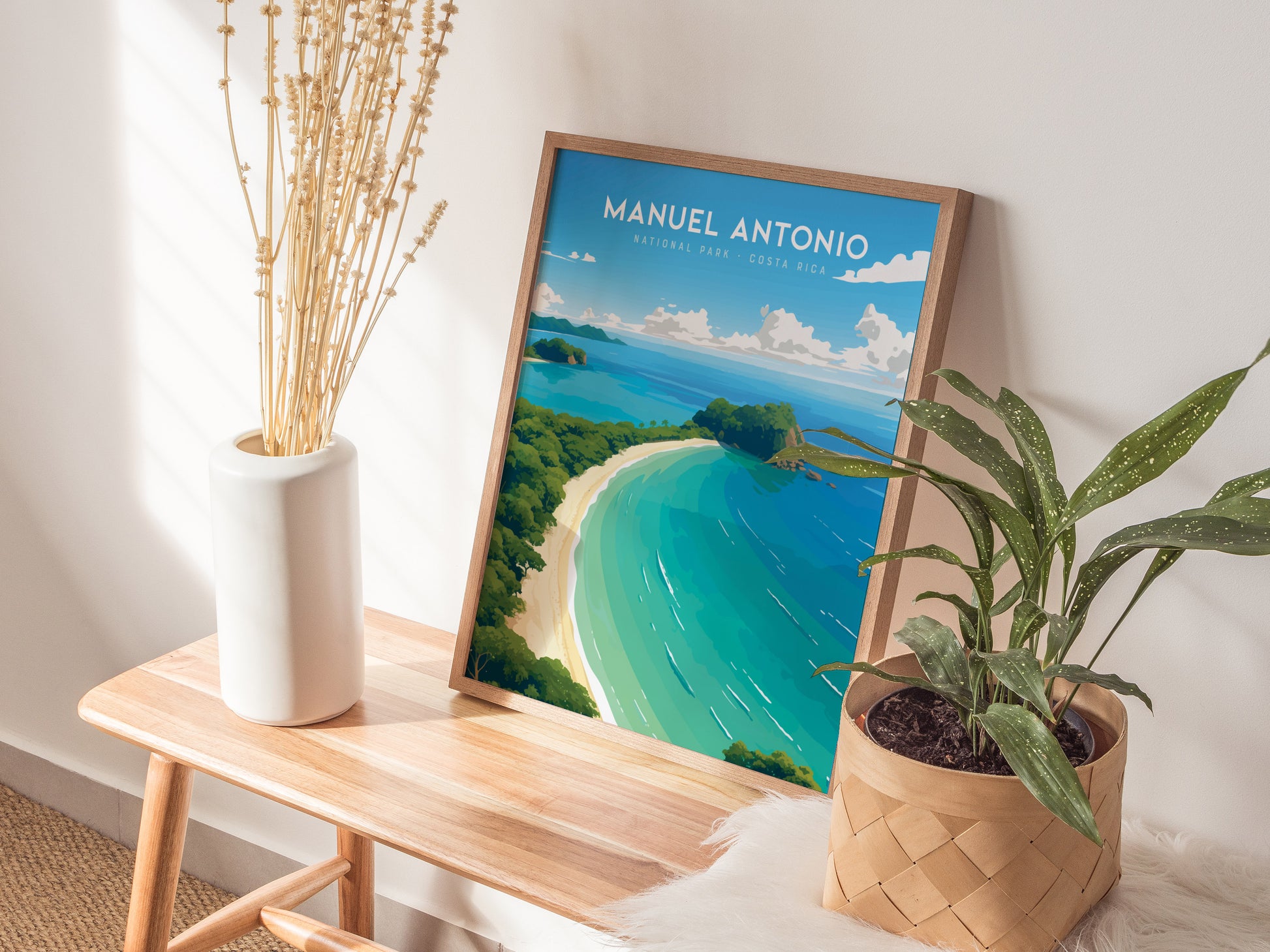 A framed poster of Manuel Antonio National Park rests on a wooden bench, surrounded by a white vase with dried flowers and a potted green plant. Sunlight casts soft shadows.