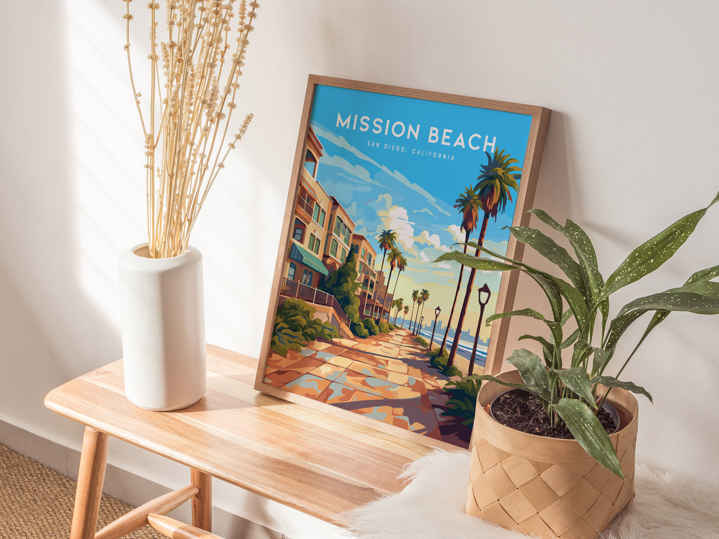 A Mission Beach San Diego poster in a wooden frame rests on a wooden bench, flanked by a potted plant and a white vase with dried branches.