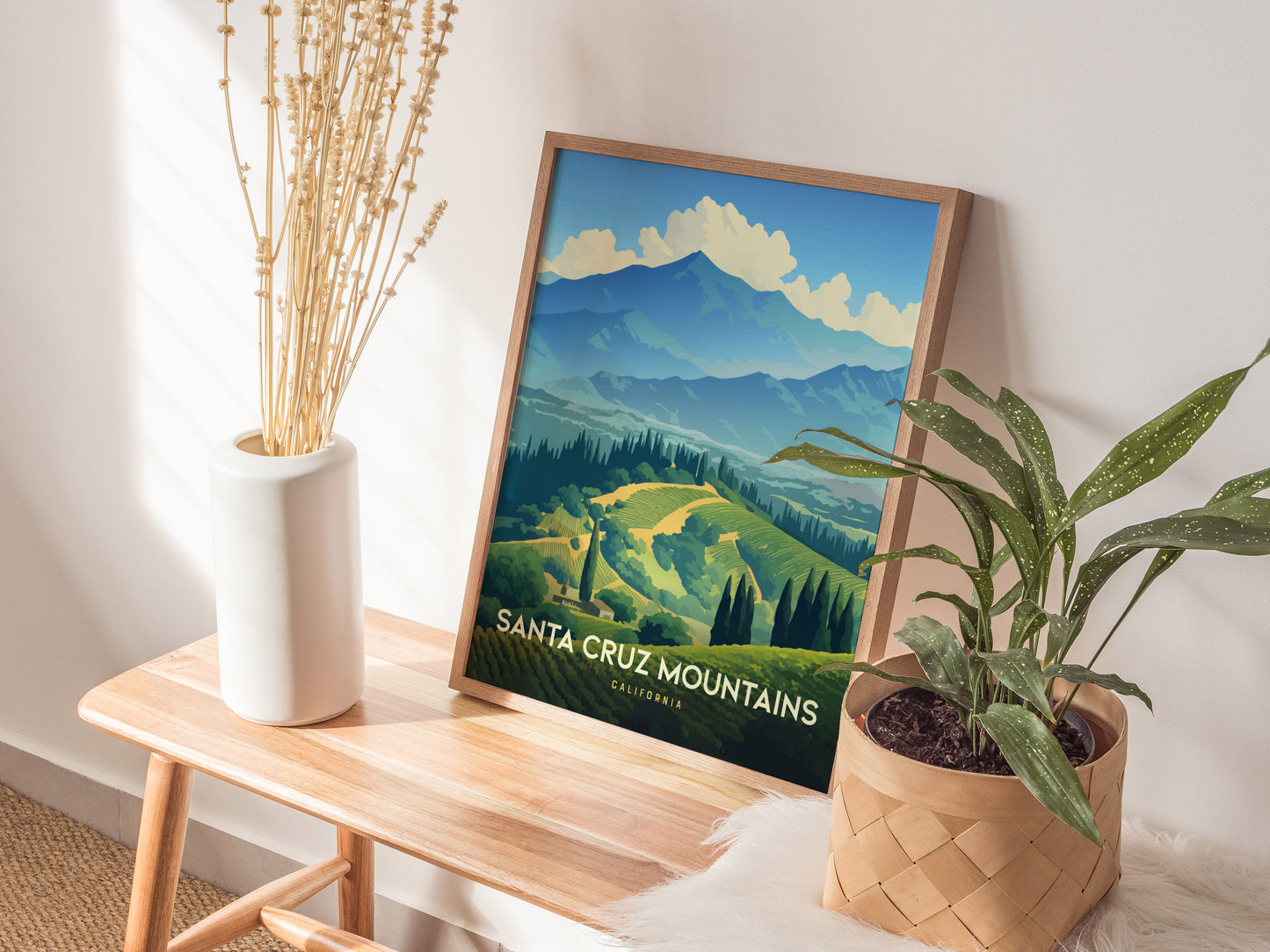 A framed Santa Cruz Mountains poster rests on a wooden bench beside a white vase with dried stems and a potted green plant. Sunlight casts soft shadows on the wall.