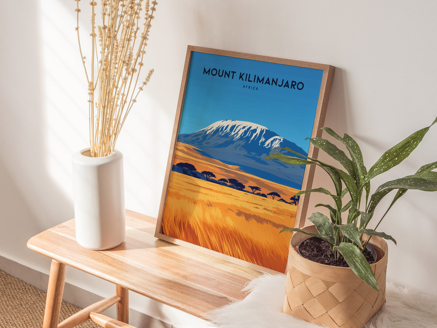 A Mount Kilimanjaro poster in a wooden frame rests on a wooden bench, flanked by a white vase with dried flowers and a potted plant.