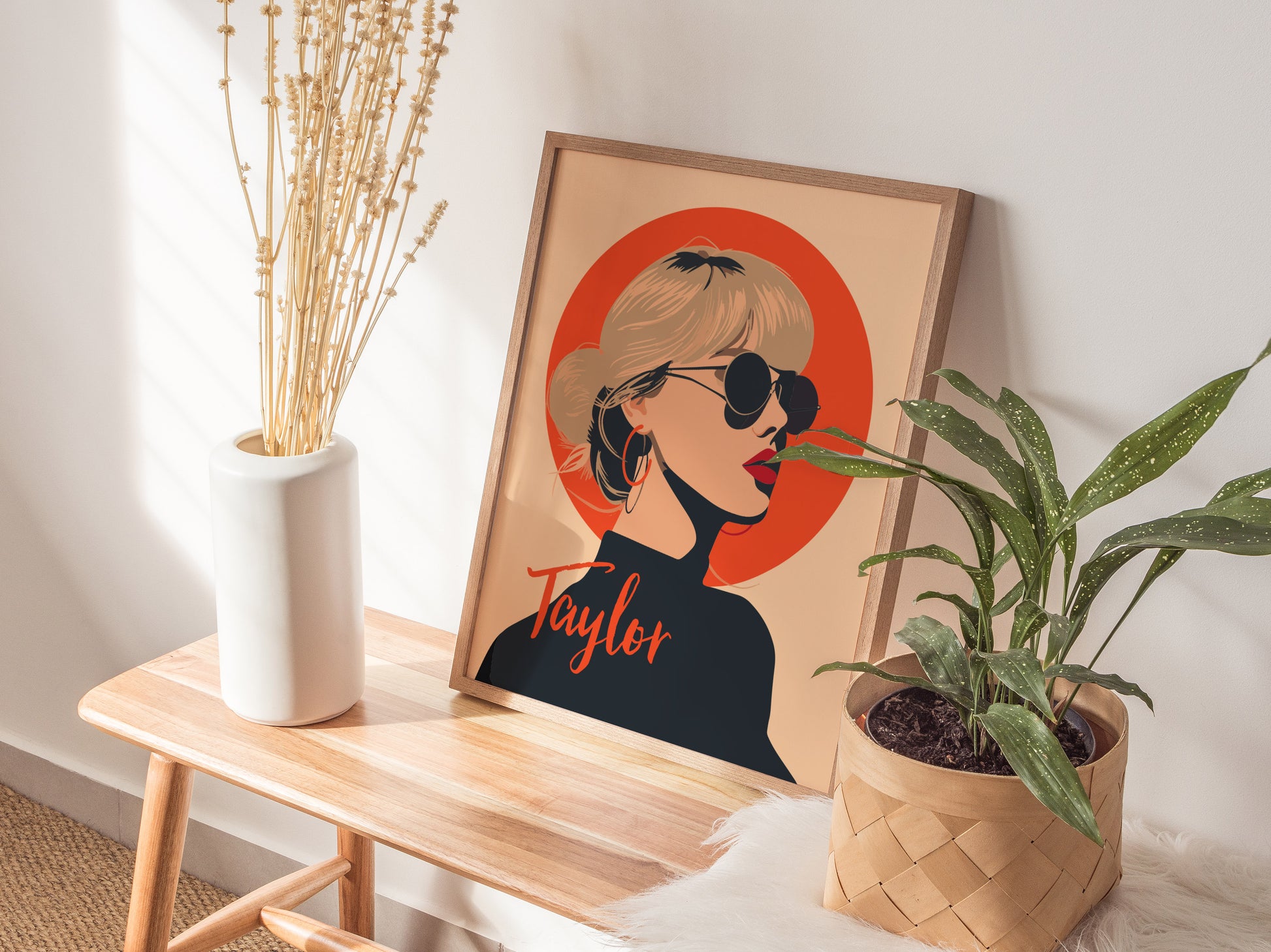 Framed minimalist poster of a woman in sunglasses with an orange background, placed on a wooden bench beside a white vase with dried stems and a potted plant.