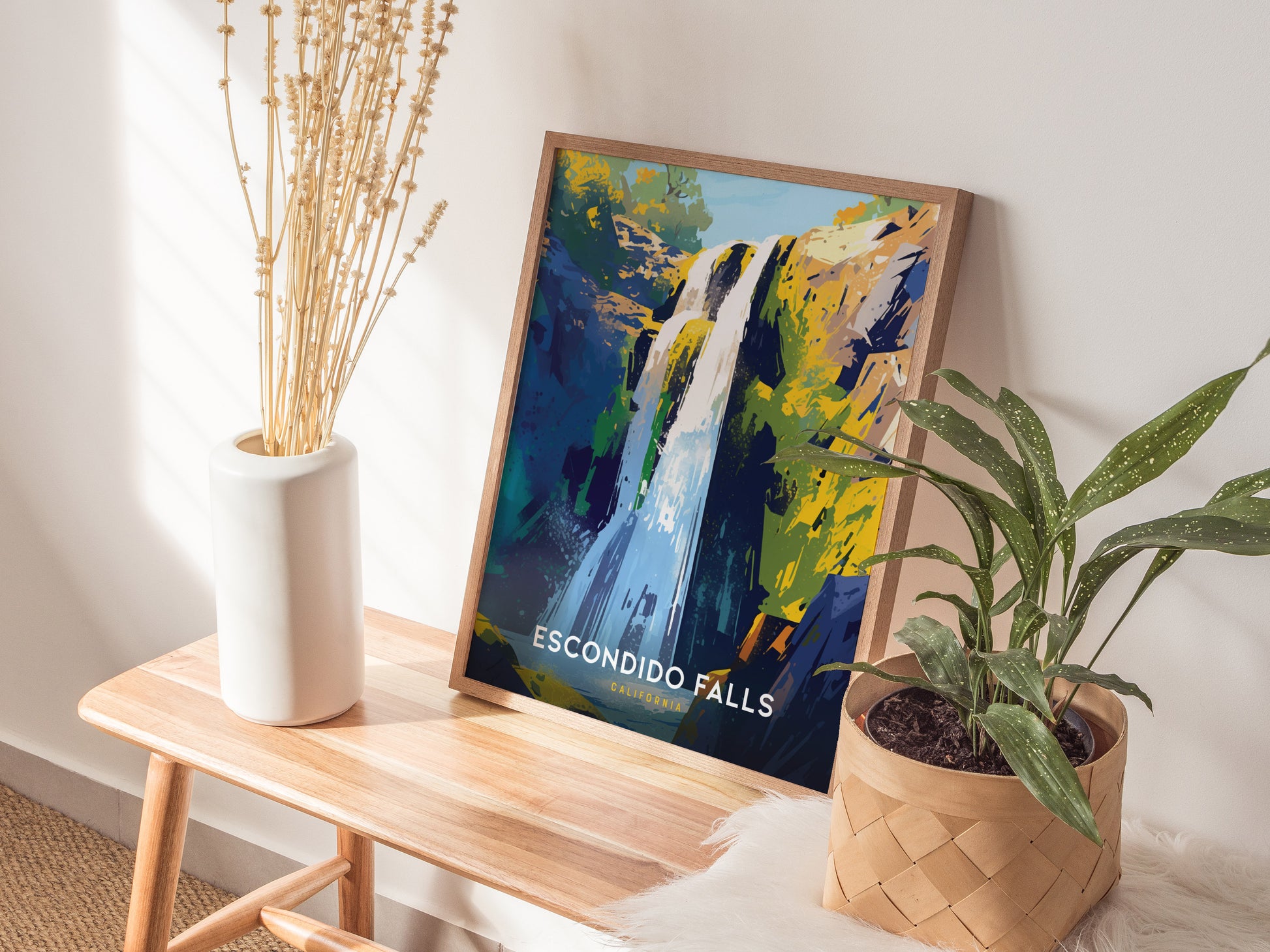 A colorful Escondido Falls poster is displayed on a wooden bench beside a white vase with dried flowers and a green potted plant. Sunlight casts shadows on the wall.