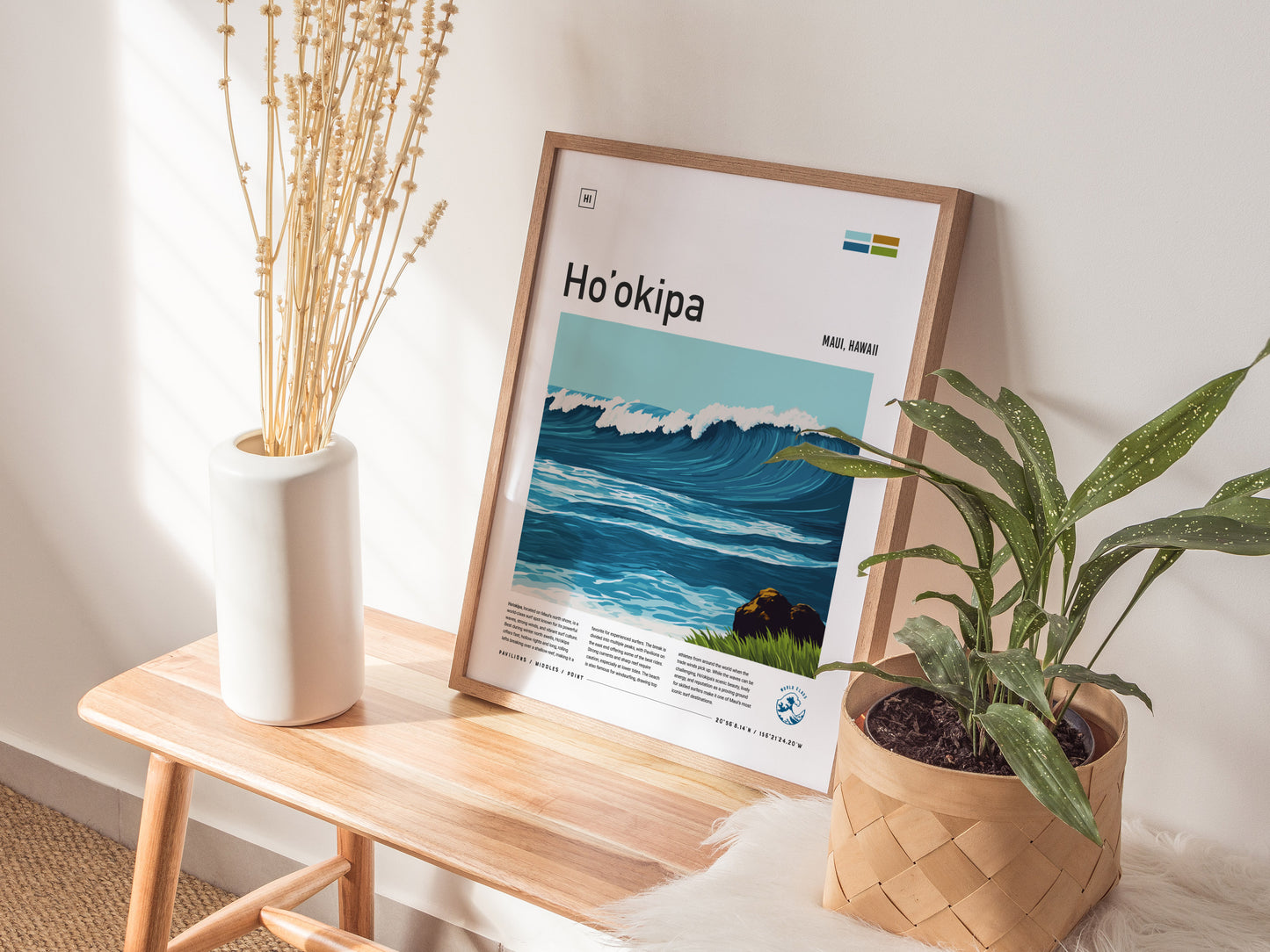 A framed Hookipa Beach poster featuring ocean waves is displayed on a wooden bench, accompanied by a white vase with dried branches and a potted green plant.