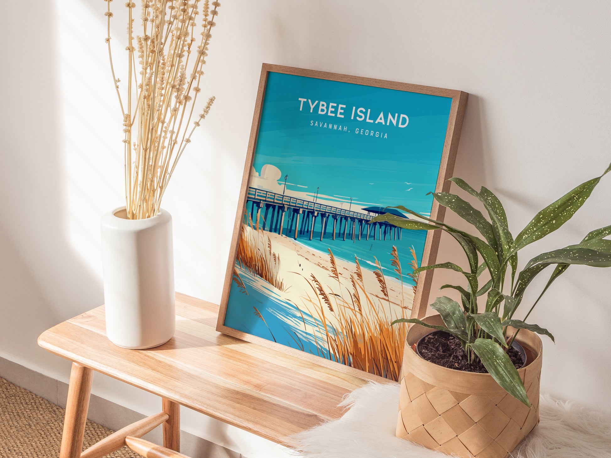 A framed Tybee Island poster featuring a beach scene with a pier and sea oats rests on a wooden bench beside a white vase with tall dried flowers and a.