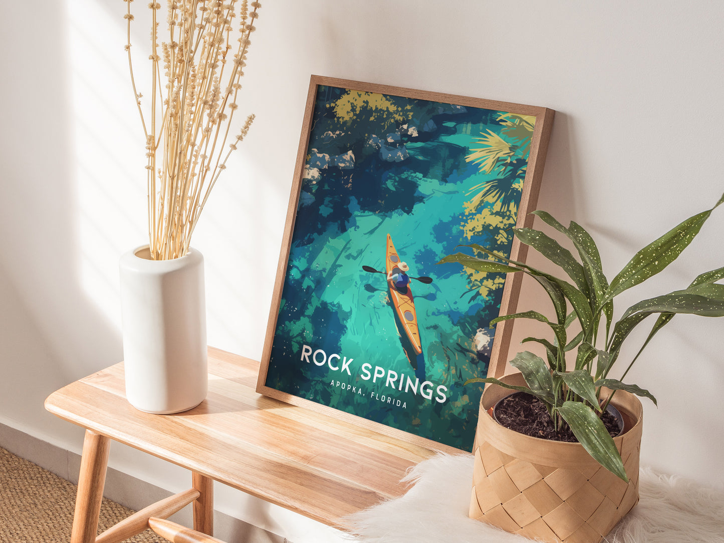 A framed Rock Springs, Apopka Florida poster featuring a kayaker on blue waters is displayed on a wooden bench next to a white vase with dried flowers and a green.