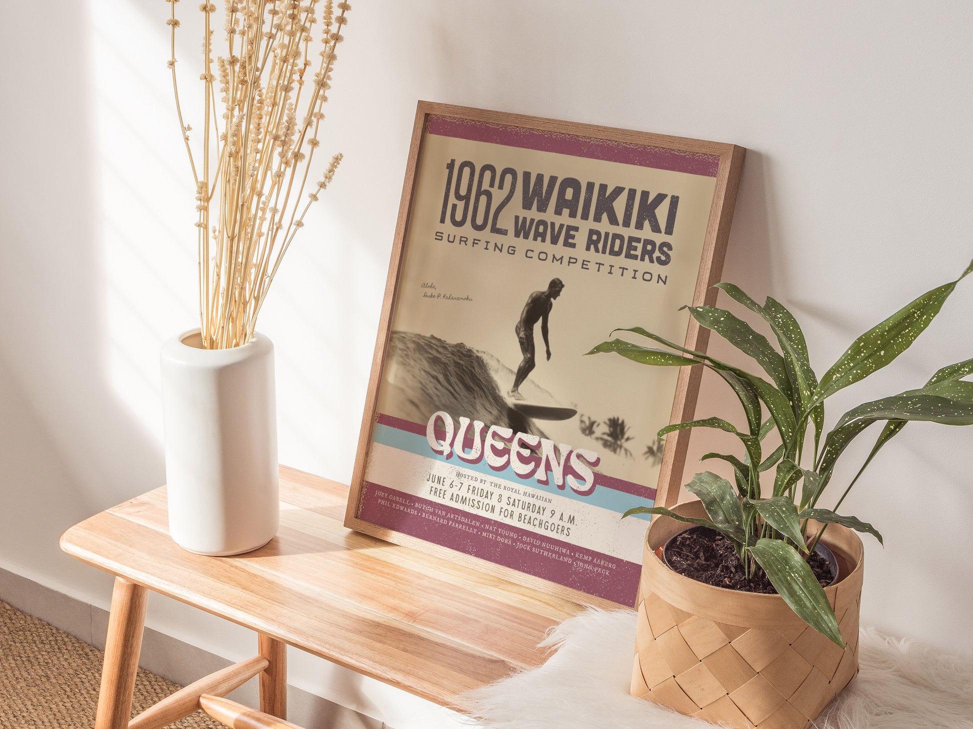 A framed 1962 Waikiki Wave Riders poster sits on a wooden bench beside a white vase with dried stems and a green potted plant. Sunlight casts soft shadows.