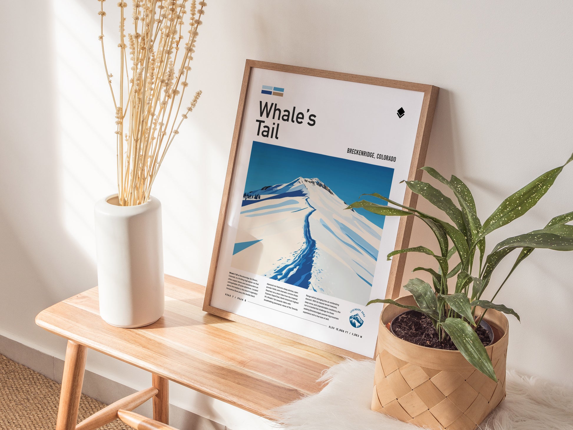 A framed poster titled Whales Tail featuring a snowy mountain scene is displayed on a wooden bench, accompanied by a white vase with dried flowers and a potted plant.