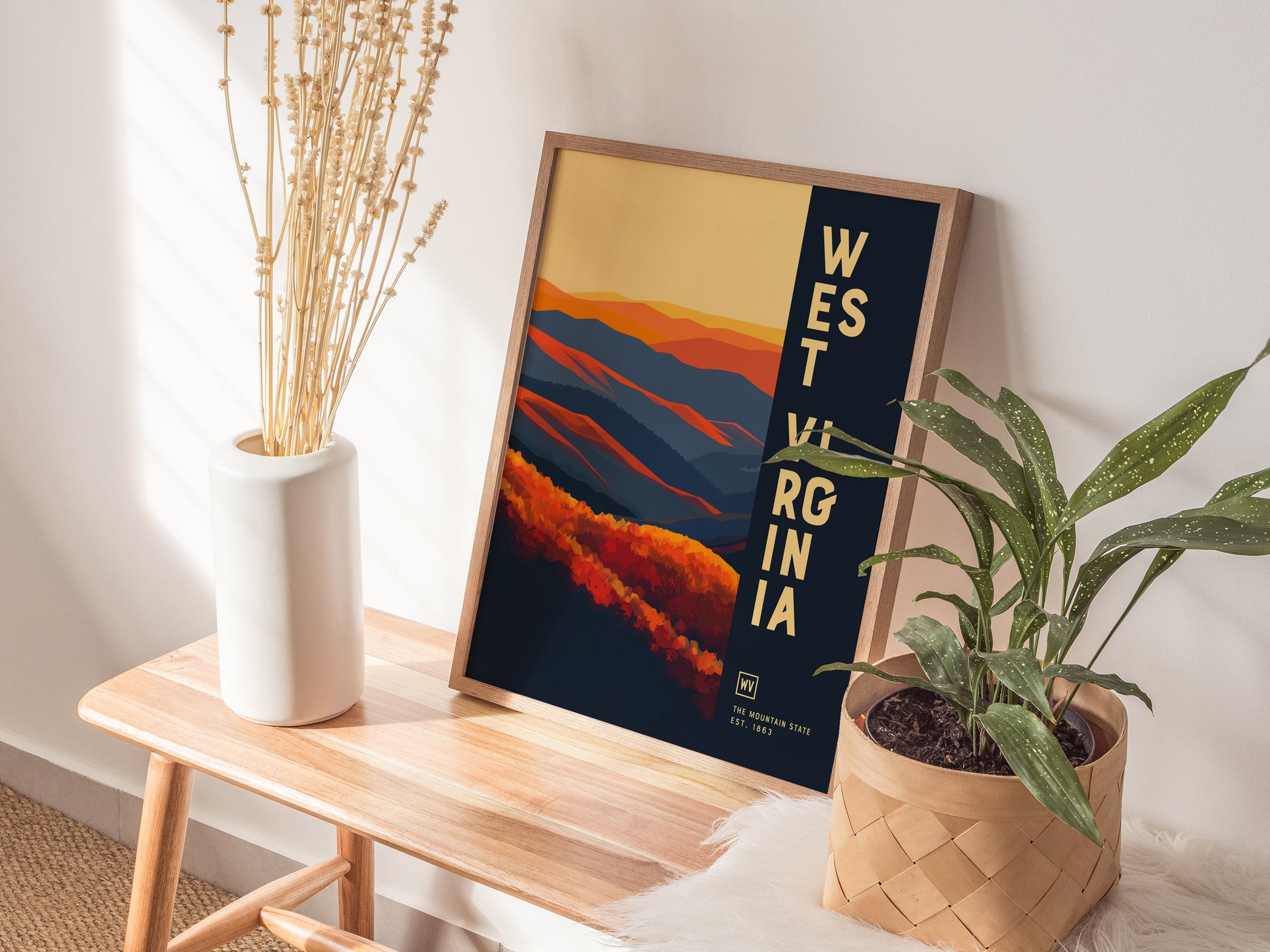 A framed West Virginia poster with vibrant abstract mountain scenery in orange and blue tones is displayed on a wooden bench next to a white vase with dried flowers and.
