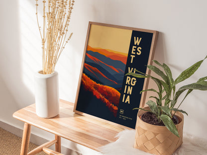 A framed West Virginia poster with vibrant abstract mountain scenery in orange and blue tones is displayed on a wooden bench next to a white vase with dried flowers and.