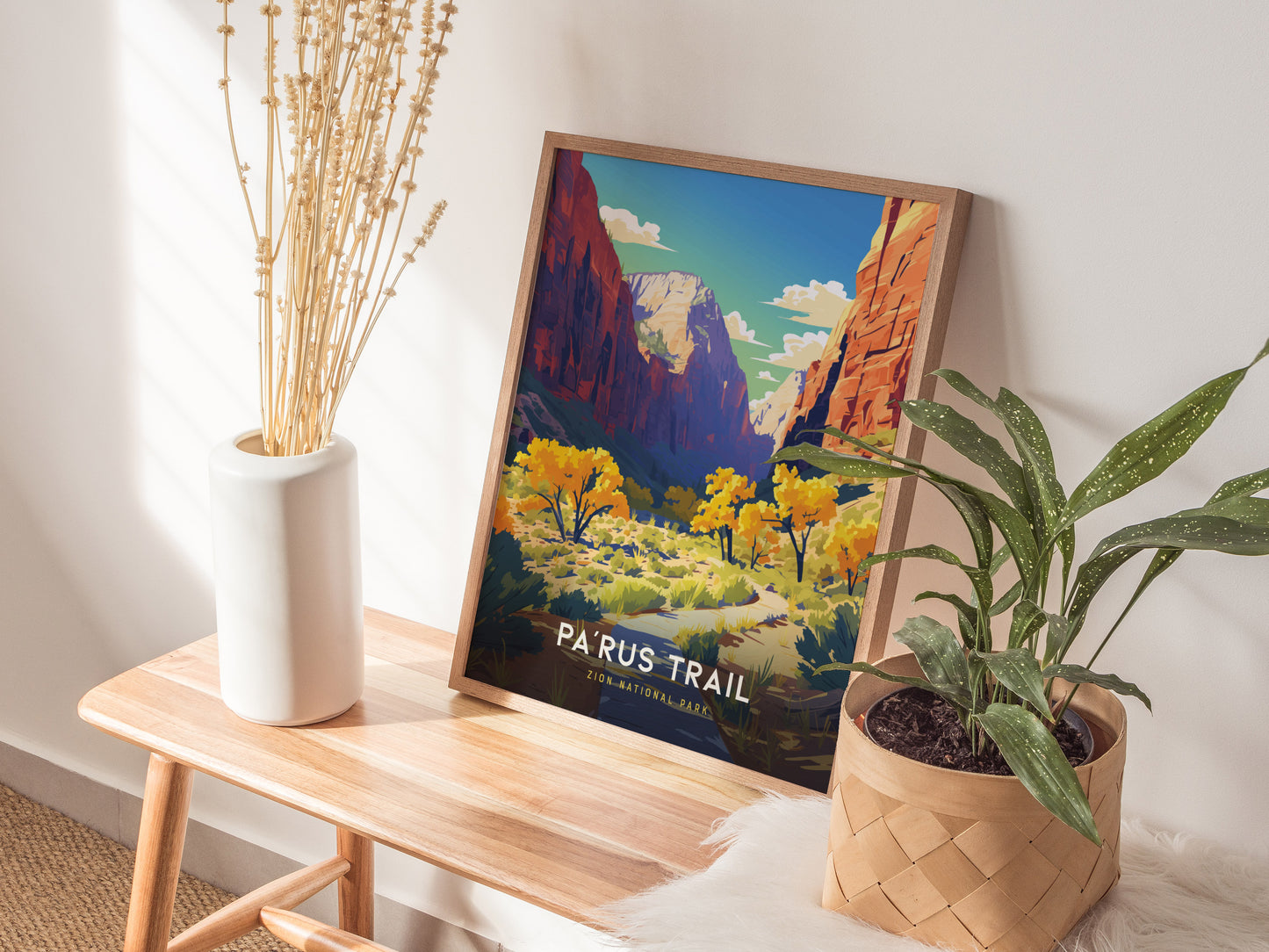 A framed poster of Zion National Parks Parus Trail rests on a wooden bench, accompanied by a white vase with dried flowers and a potted green plant.