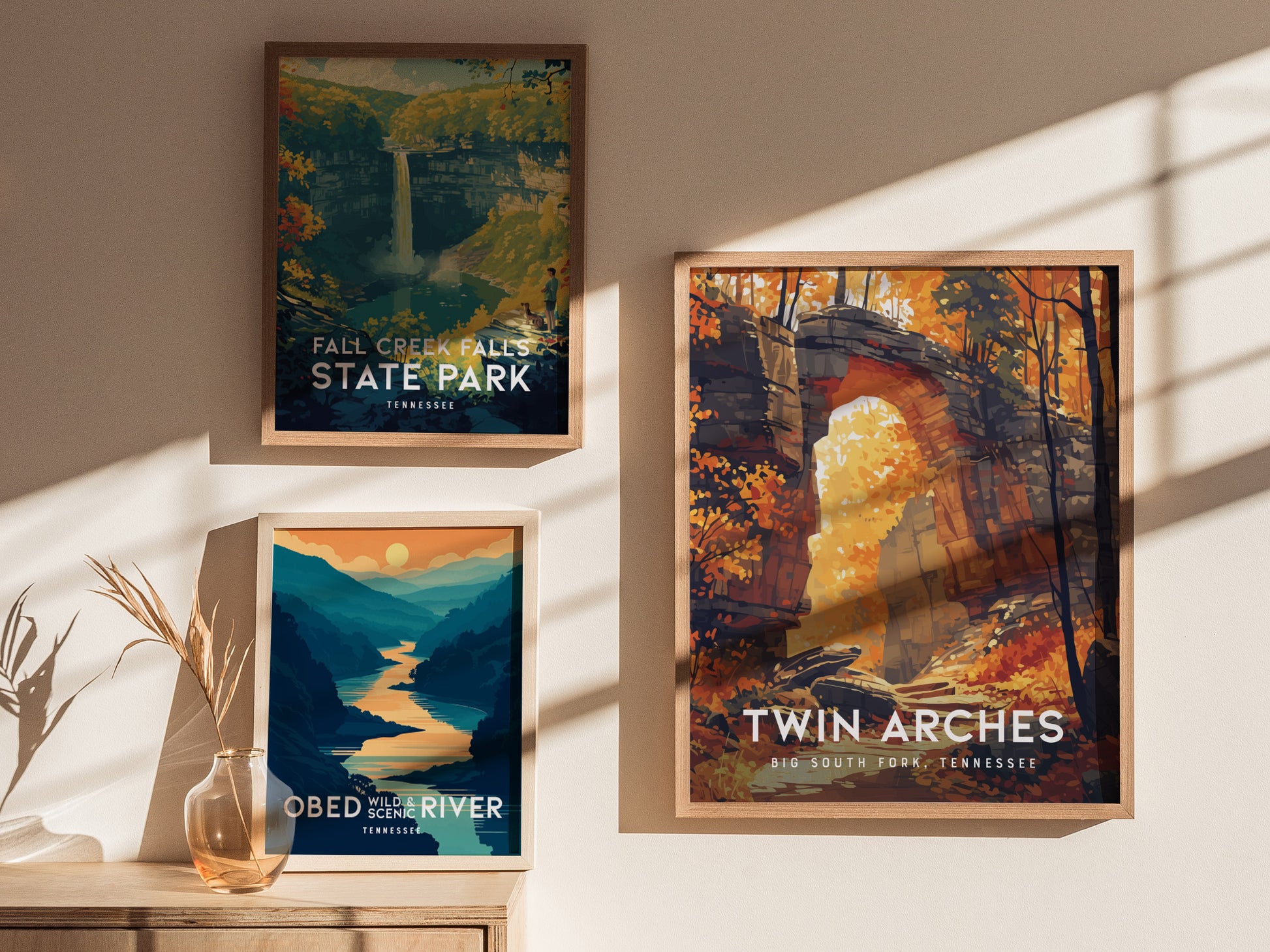 Three framed posters on a wall show stylized landscapes: Fall Creek Falls, Obed Wild and Scenic River, and Twin Arches in autumn. Sunlight casts shadows across the wall.