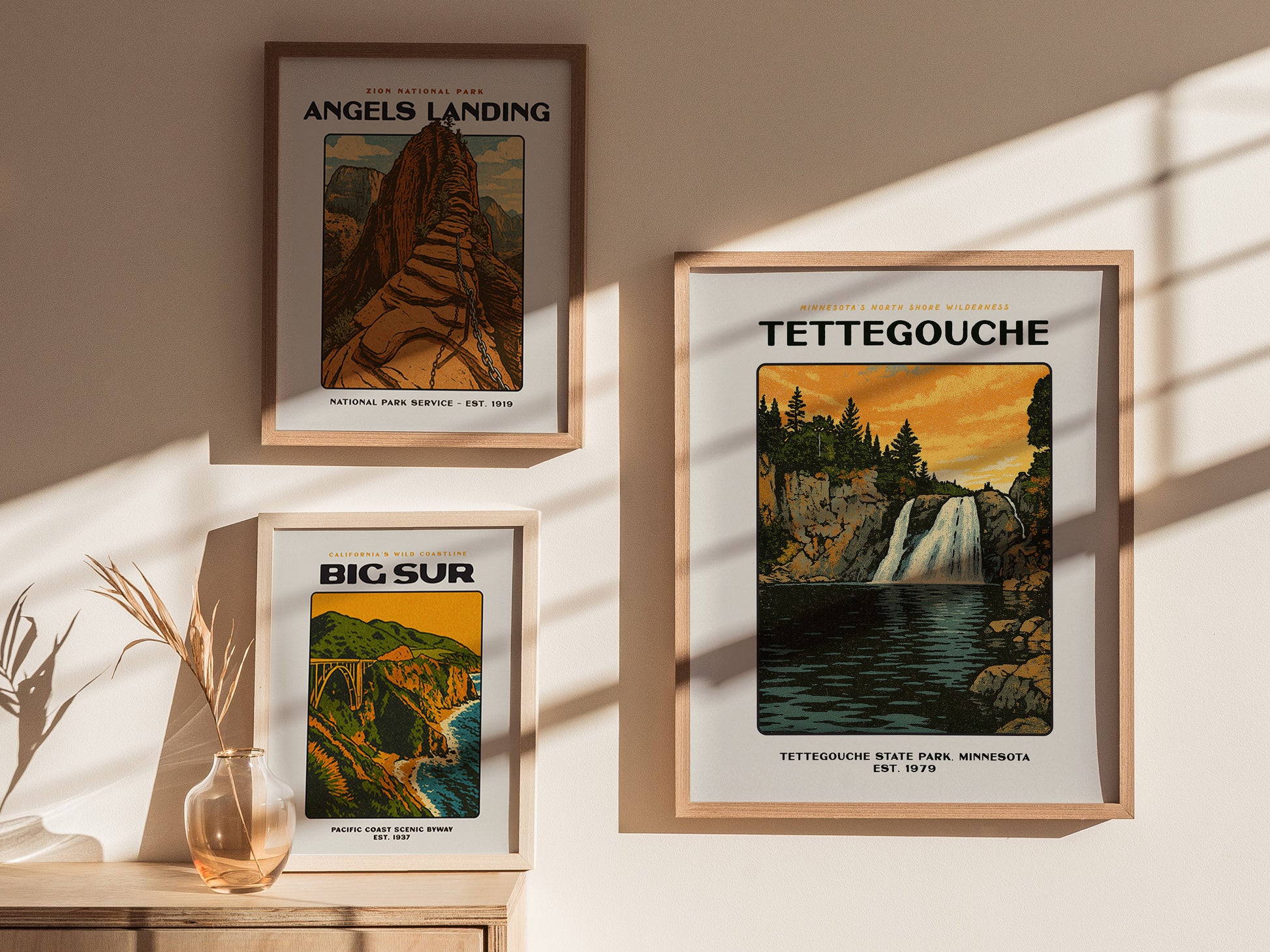Three framed posters are displayed on a sunlit wall: Angels Landing, Big Sur, and Tettegouche featuring a waterfall. A vase with dried plants is on a wooden surface.
