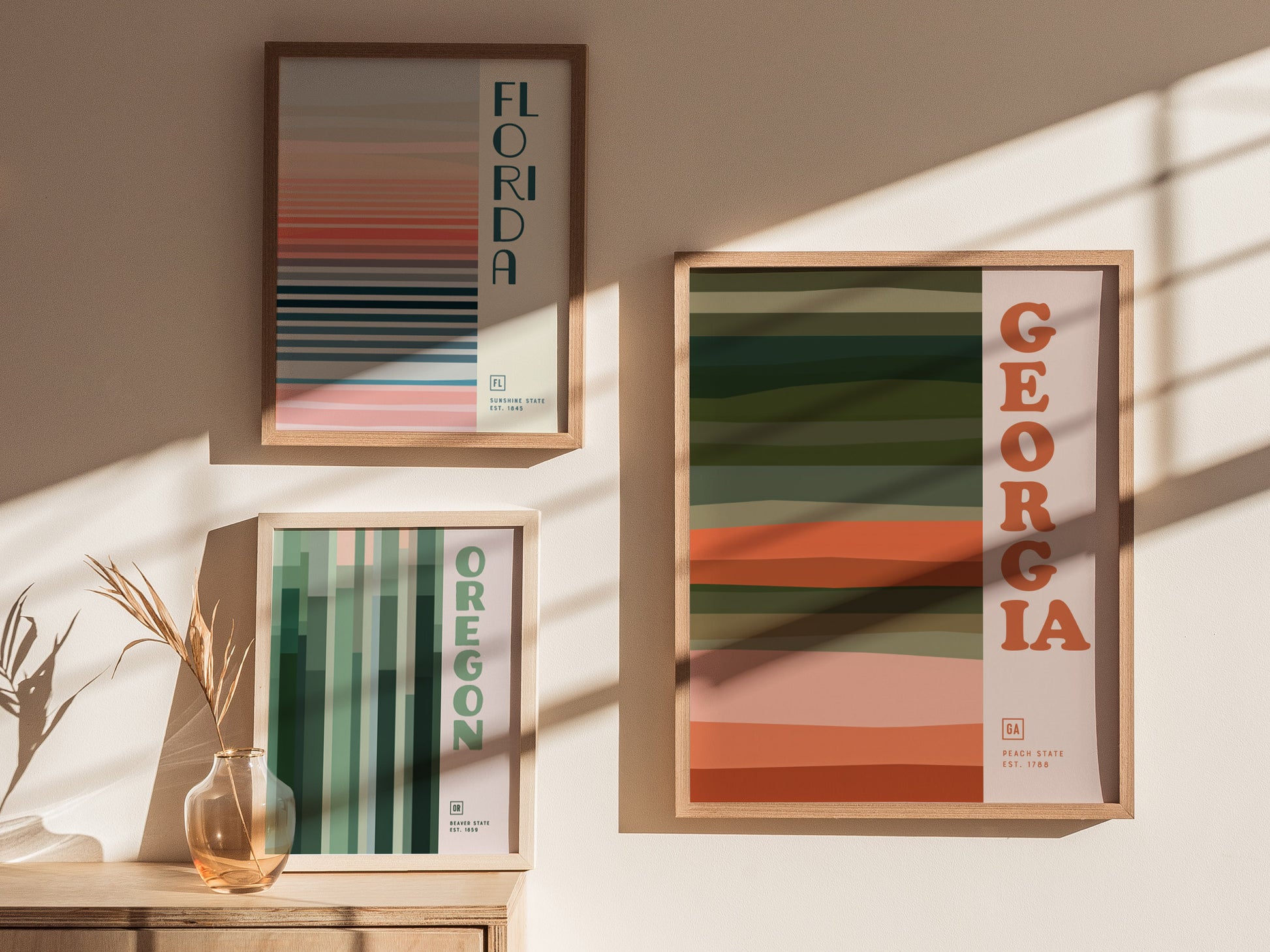 Three framed posters featuring minimalist state designs for Florida, Oregon, and Georgia with vertical and horizontal stripes in earth tones, displayed on a sunlit wall.