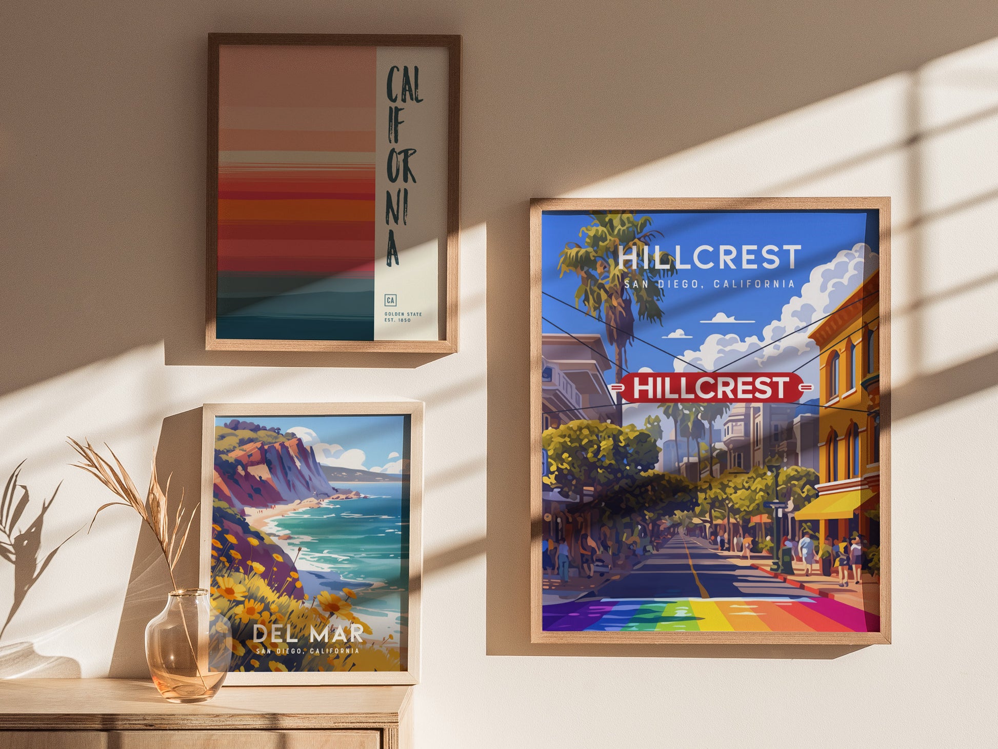 Framed posters on a wall feature vibrant illustrations of Hillcrest, Del Mar, and a California-themed design. Sunlight casts shadows on the wall and a table with a vase.