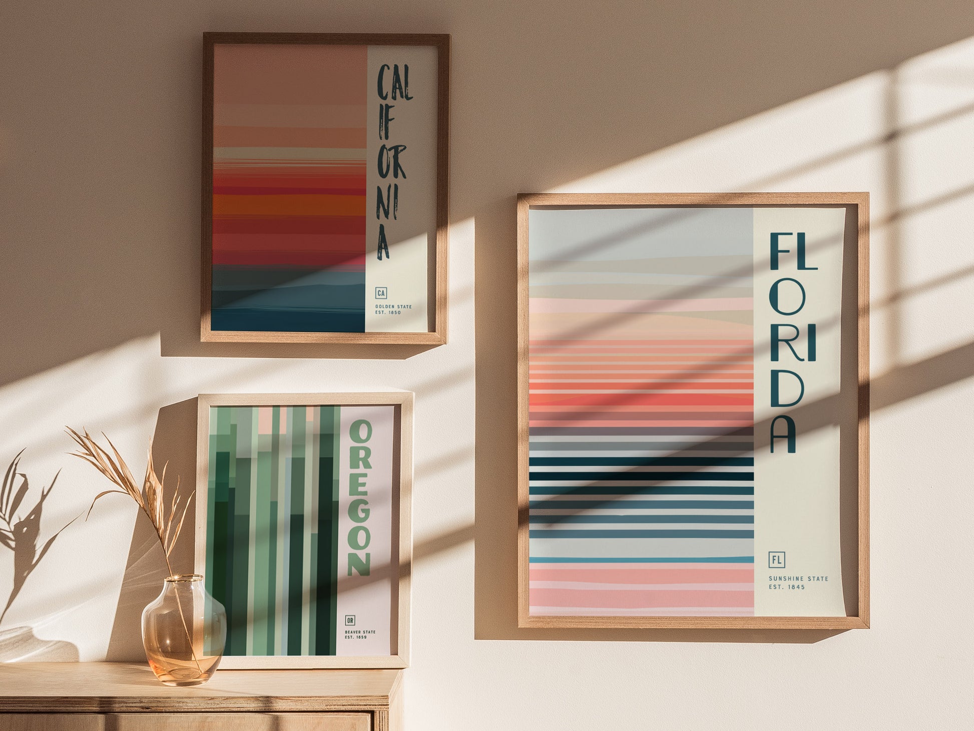 Three minimalist posters featuring California, Oregon, and Florida designs hang on a sunlit wall. Each poster displays vertical or horizontal stripes in varying pastel colors.