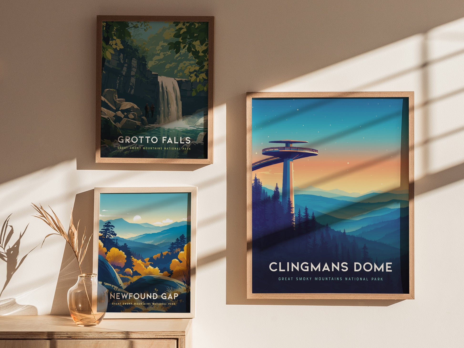 Three framed posters are displayed on a sunlit wall. The top left features Grotto Falls with a waterfall scene, the bottom left shows Newfound Gap with autumn foliage, and the right poster depicts Clingmans Dome at sunset with rolling.
