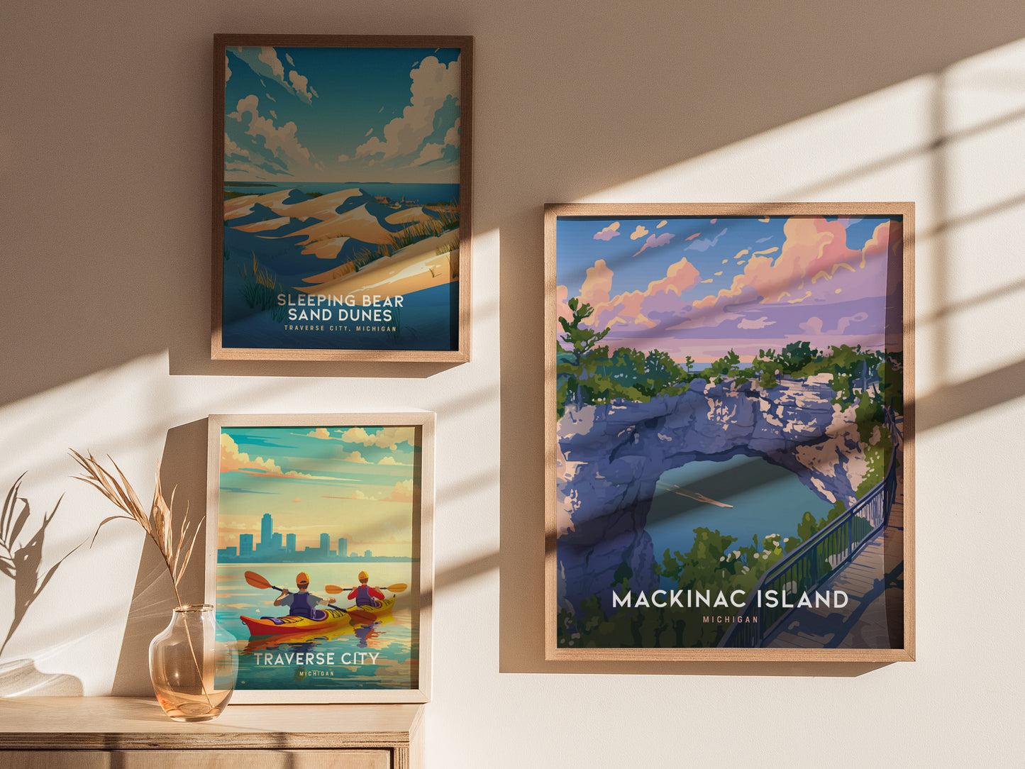 Three framed posters hang on a sunlit wall. They depict Sleeping Bear Sand Dunes, Traverse City with kayakers, and Mackinac Islands Arch Rock with vibrant colors and natural landscapes.