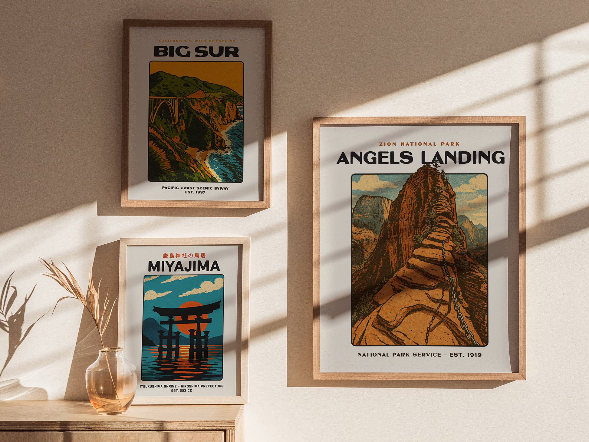 Three framed vintage-style posters are displayed on a sunlit wall, featuring Angels Landing, Big Sur, and Miyajima, with a decorative vase and dried plant on a wooden surface.