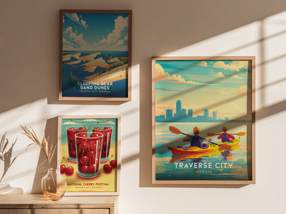 Three framed posters are displayed on a wall, featuring Sleeping Bear Sand Dunes, a National Cherry Festival, and kayakers on Grand Traverse Bay. Sunlight casts shadows across the scene.