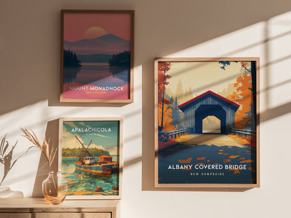 Three framed posters are displayed on a wall: one features the Albany Covered Bridge in New Hampshire with autumn foliage, another depicts Mount Monadnock, and the third shows a colorful boat scene in Apalachicola.