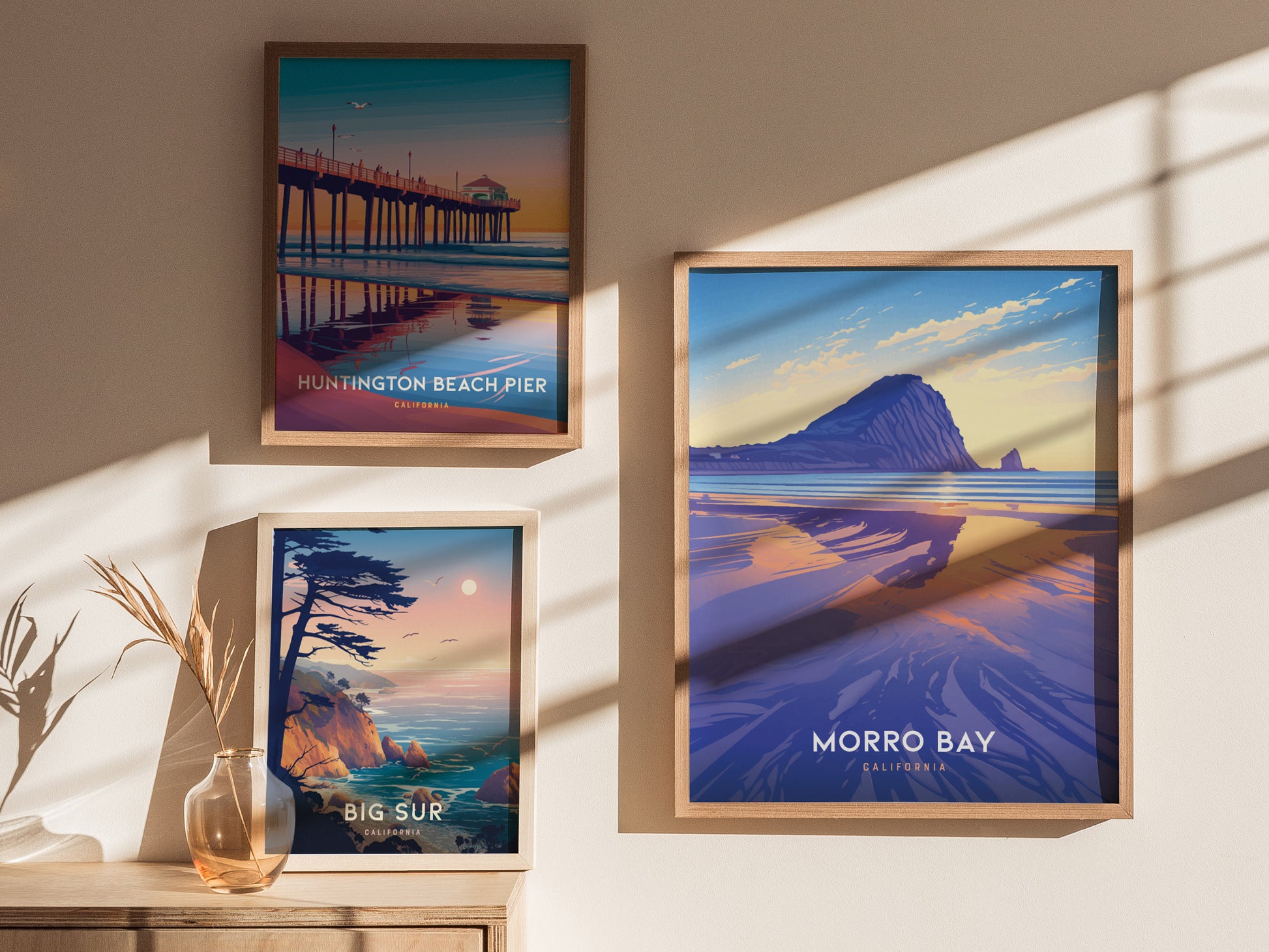 Three framed posters on a wall depict scenic California locations: Huntington Beach Pier, Big Sur, and Morro Bay, with natural lighting casting shadows across the artwork.