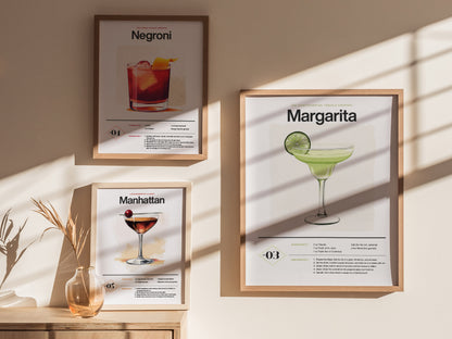 Three framed cocktail posters are displayed on a wall, featuring Negroni, Manhattan, and Margarita drinks. Sunlight casts shadows, and a glass vase with dried grass is nearby.
