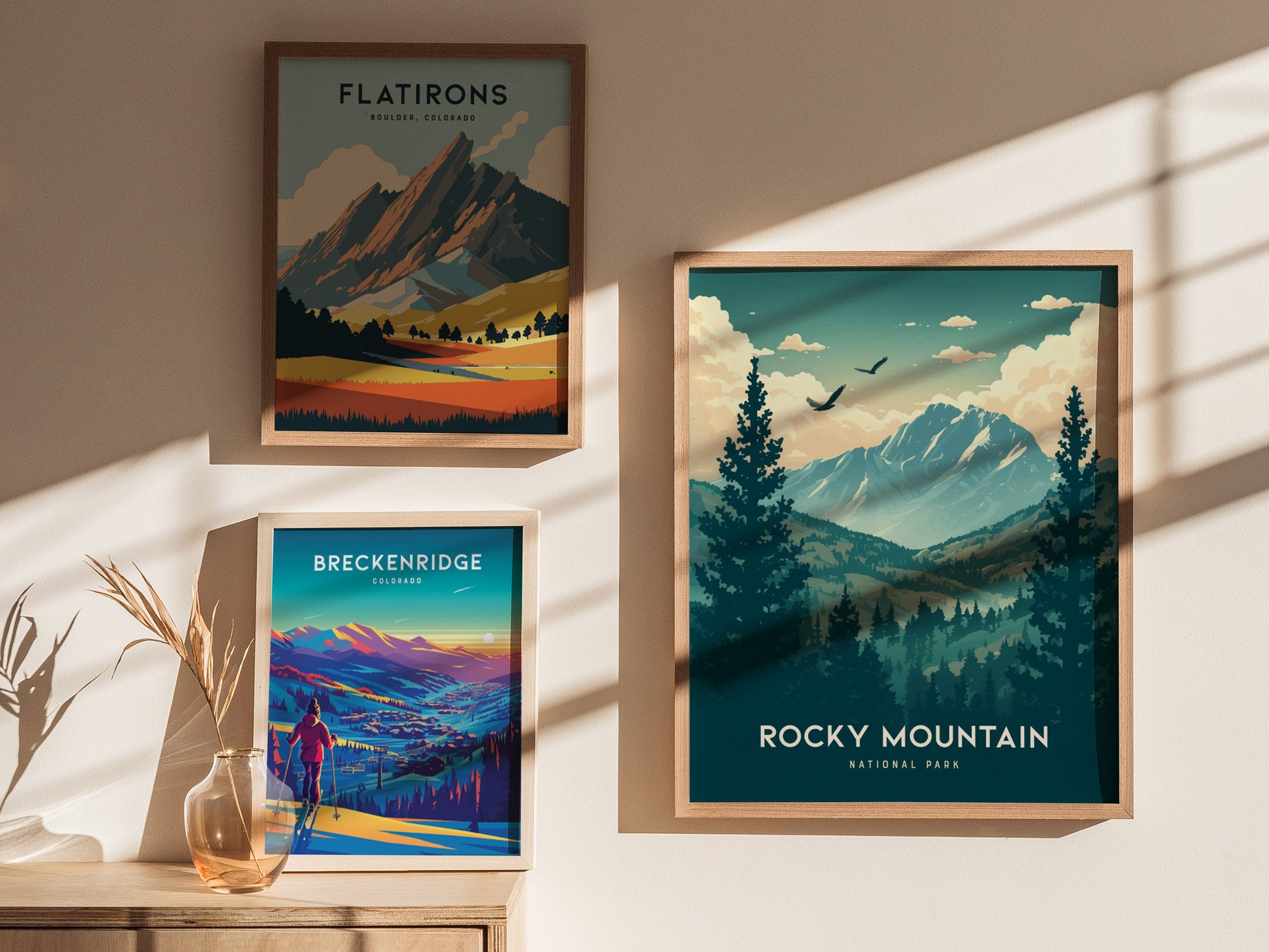 Framed posters of Flatirons, Breckenridge, and Rocky Mountain National Park hang on a sunlit wall. A vase with dried grasses sits on a wooden surface beneath them.