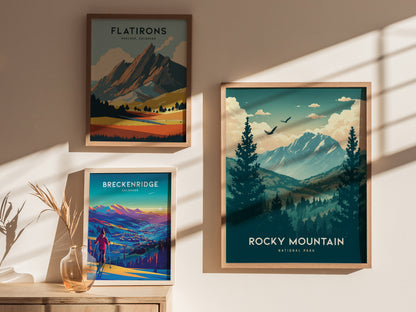 Framed posters of Flatirons, Breckenridge, and Rocky Mountain National Park hang on a sunlit wall. A vase with dried grasses sits on a wooden surface beneath them.