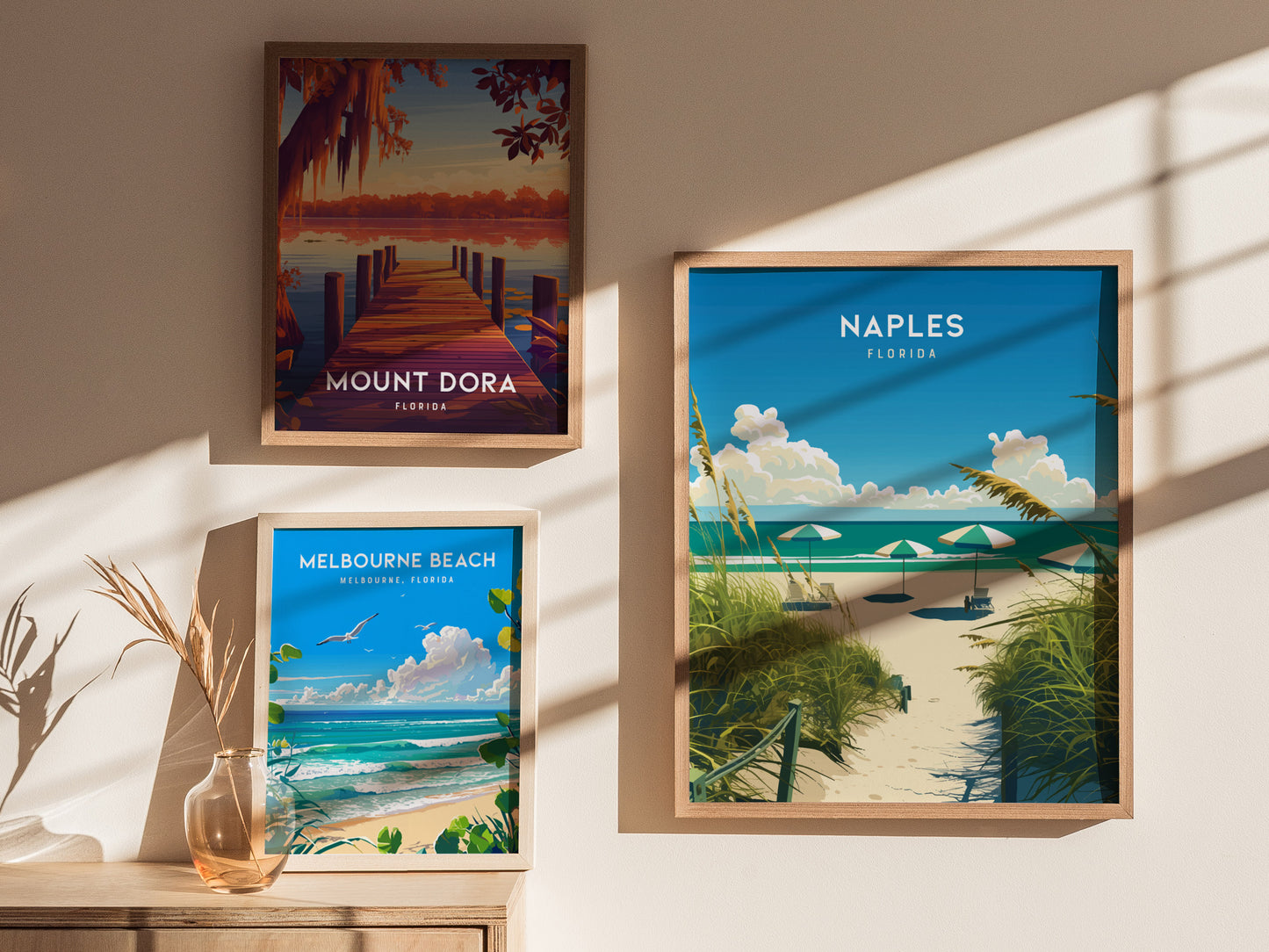 Three framed posters on a sunlit wall depict Mount Dora, Melbourne Beach, and Naples, Florida, showcasing scenic landscapes with water, foliage, and vibrant skies.