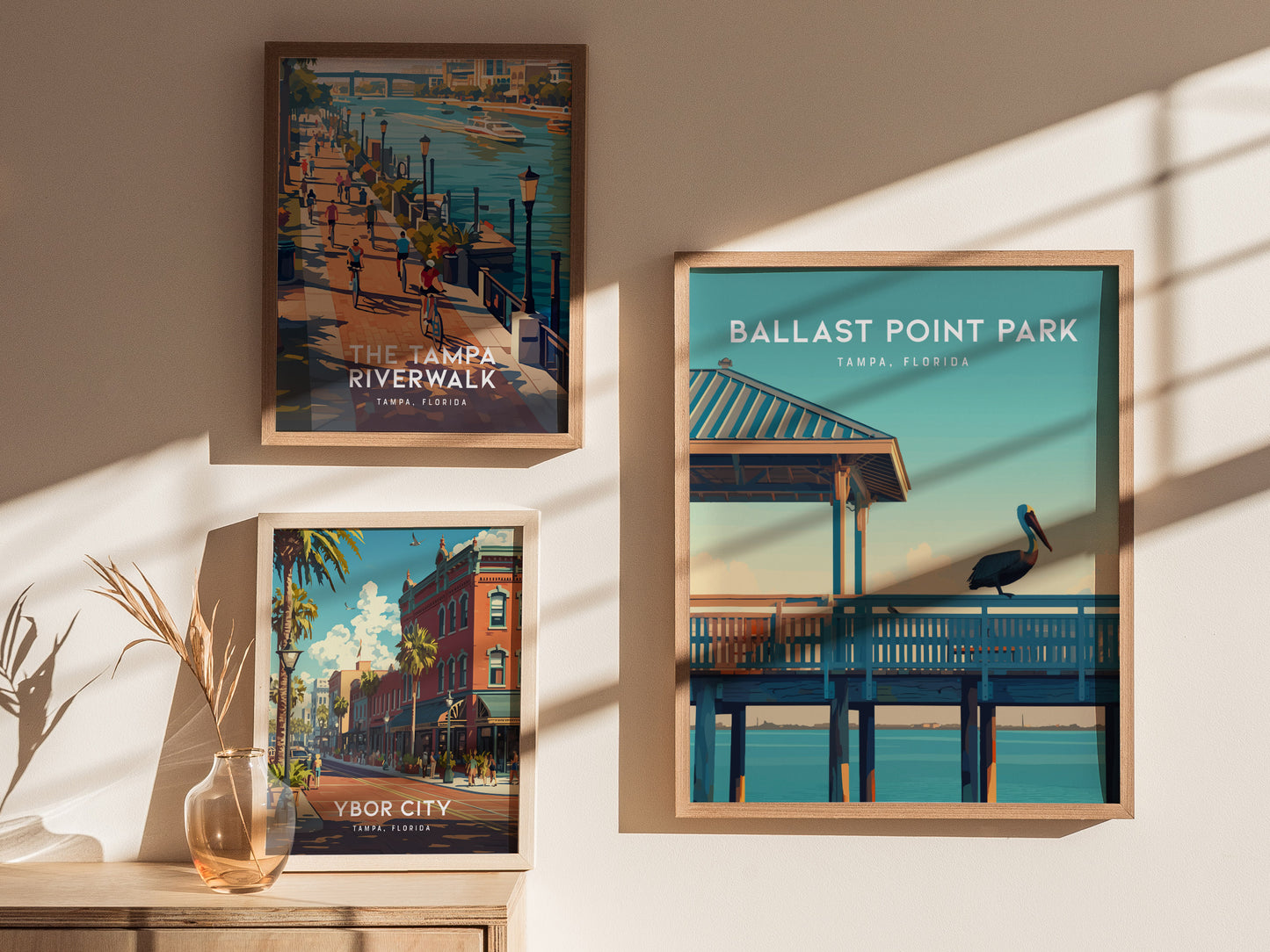 Three framed posters are displayed on a wall, featuring scenic illustrations of Tampa Riverwalk, Ballast Point Park with a pelican, and Ybor City, accompanied by a vase with dried plants.