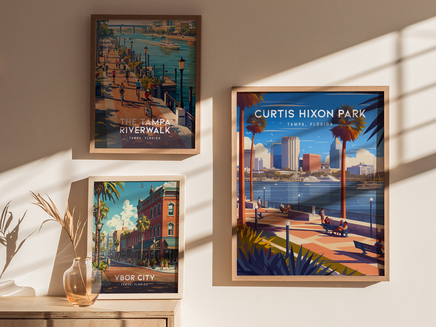 Three framed posters on a wall depict Tampa, Florida scenes, including Curtis Hixon Park, a riverwalk, and Ybor City, with a vase of dried plants nearby.