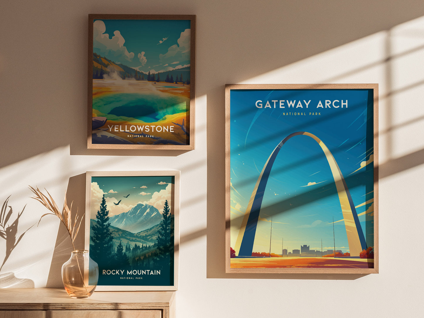 Three framed posters are displayed on a wall: Gateway Arch, Yellowstone, and Rocky Mountain, with sunlight casting shadows and a decorative vase with dried plants nearby.