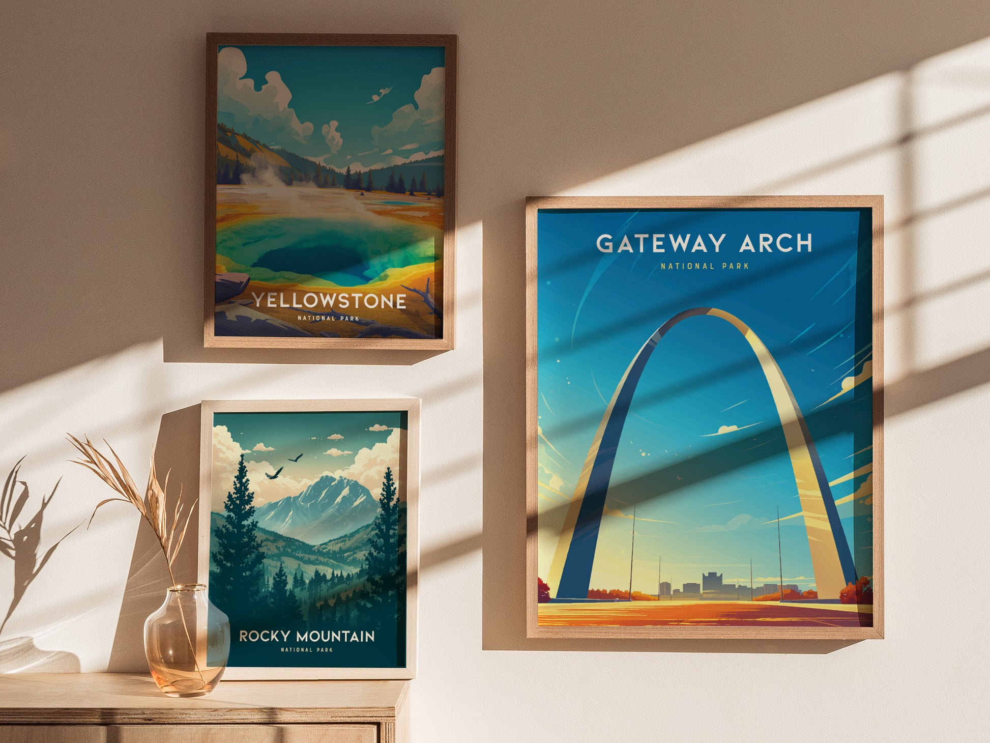 Three framed posters are displayed on a wall: Gateway Arch, Yellowstone, and Rocky Mountain, with sunlight casting shadows and a decorative vase with dried plants nearby.