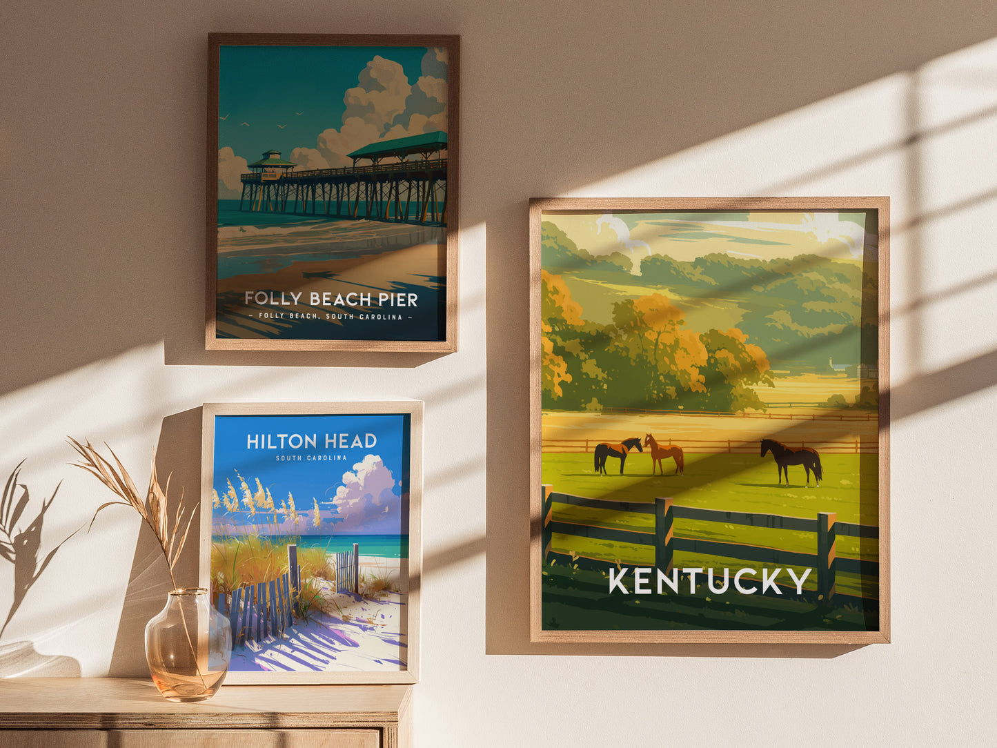 Three framed posters on a wall depict Folly Beach Pier, Hilton Head, and a Kentucky landscape with horses and a fence. Sunlight casts soft shadows on the wall.