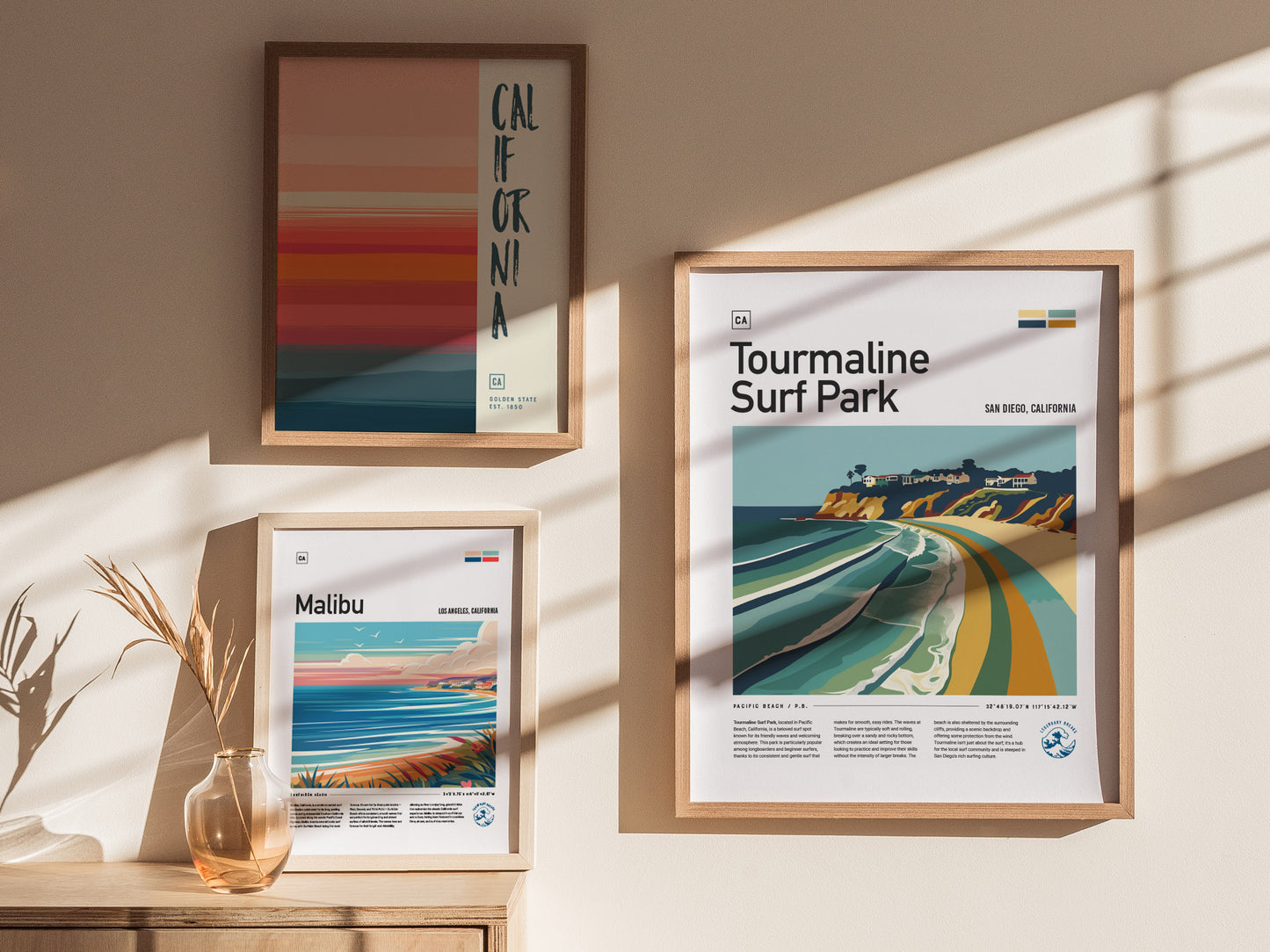 Three framed posters are displayed on a wall, featuring colorful beach art and the titles California, Tourmaline Surf Park, and Malibu, with shadows and a vase of dried grass.