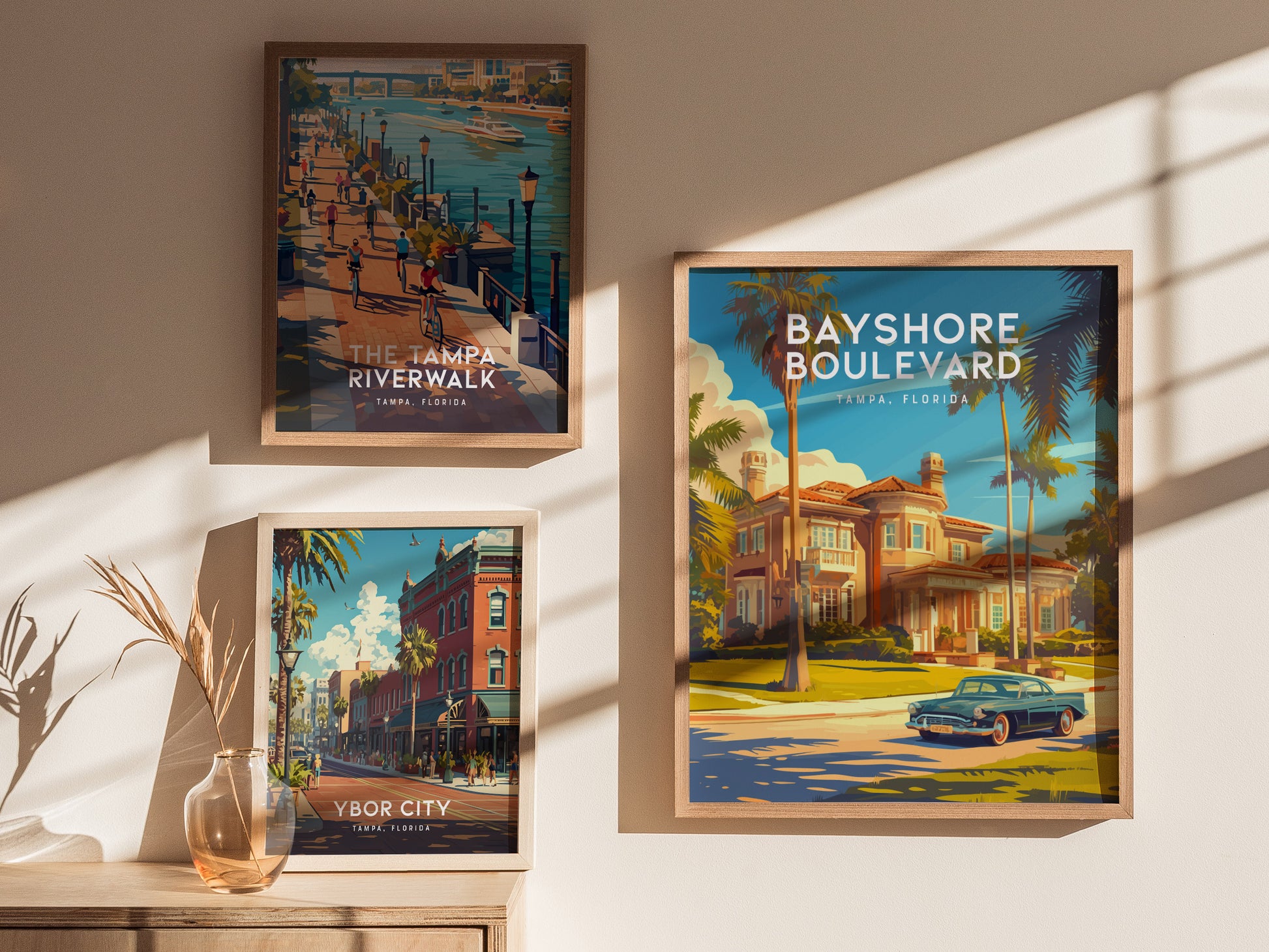 Three framed posters on a wall display scenic views of Tampa, Florida, featuring Bayshore Boulevard, the Tampa Riverwalk, and Ybor City. A vase with dry stems sits below.