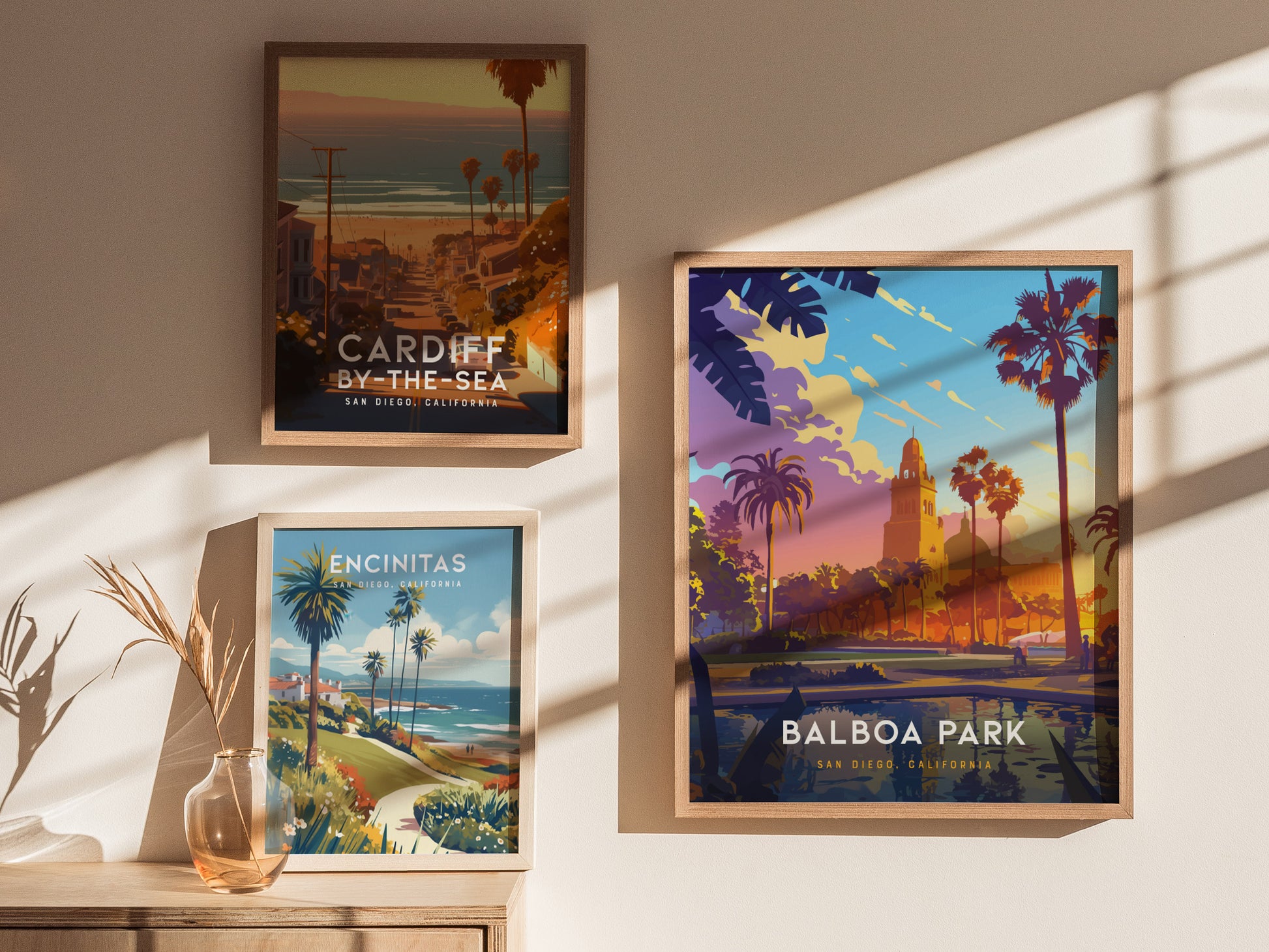 Framed posters of Cardiff-by-the-Sea, Encinitas, and Balboa Park hang on a sunlit wall, with a vase of dried plants on a wooden surface below.