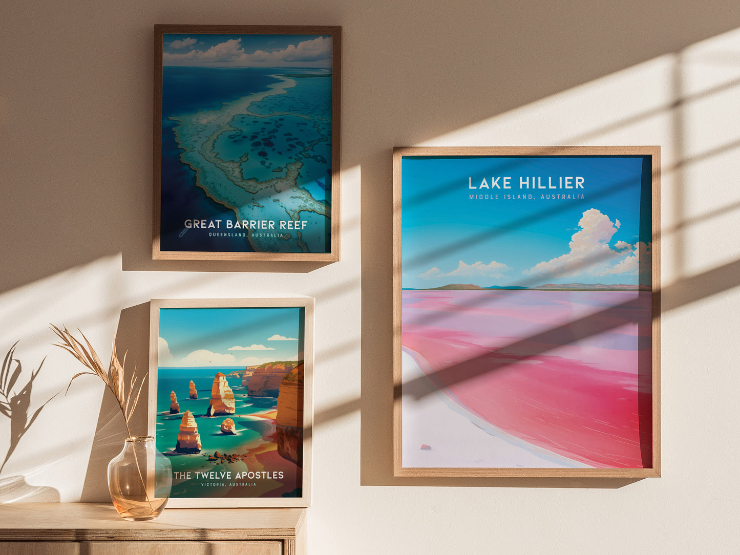 Three framed posters hang on a beige wall, featuring Lake Hillier, the Great Barrier Reef, and the Twelve Apostles. Natural light casts shadows on the wall and a table below.