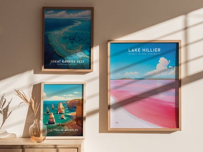 Three framed posters hang on a beige wall, featuring Lake Hillier, the Great Barrier Reef, and the Twelve Apostles. Natural light casts shadows on the wall and a table below.