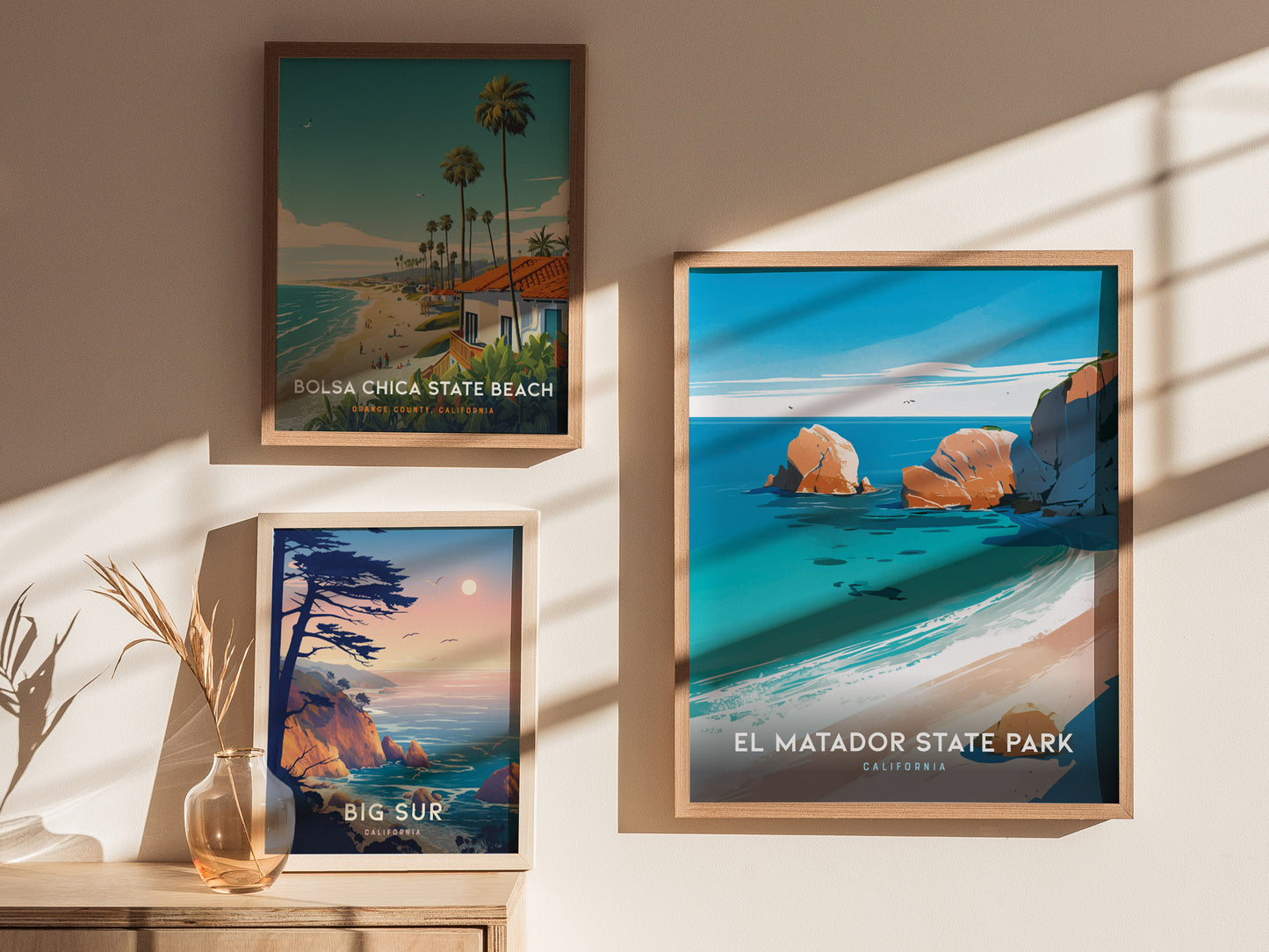 Framed posters of Bolsa Chica State Beach, Big Sur, and El Matador State Park are displayed on a wall, with sunlight casting shadows and a vase with dried plants on.