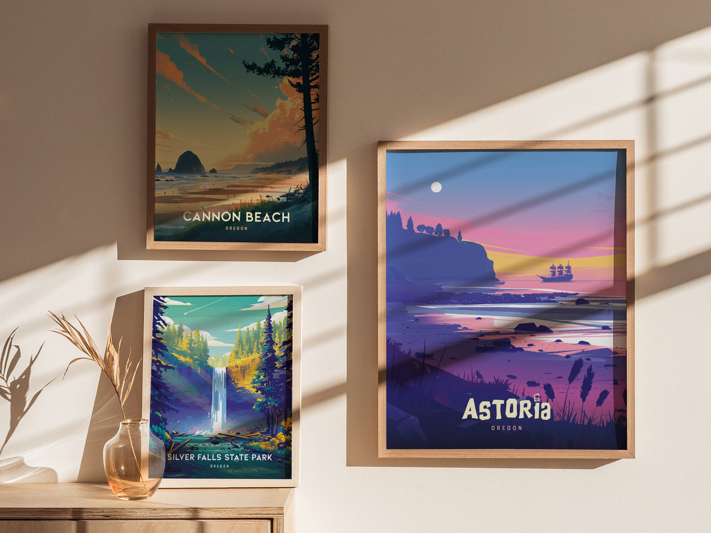 Three framed posters are displayed on a wall, featuring scenic illustrations of Cannon Beach, Silver Falls State Park, and Astoria, Oregon. A glass vase with dried grass is nearby.